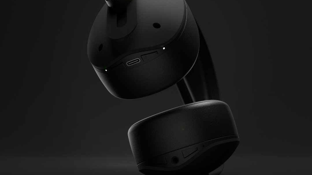 A pair of matte black over ear headphones shown with one ear cup detached, so you can see the removable pad and charging port