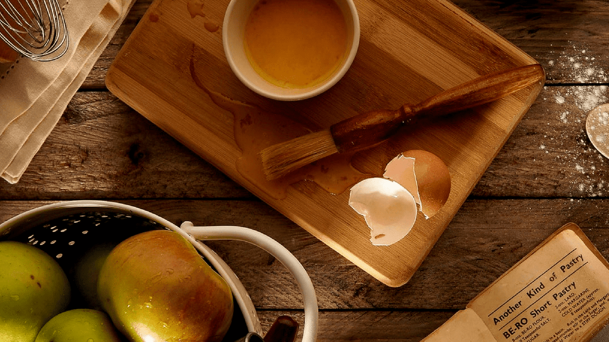 A wooden kitchen worktop laid out with a chopping board, a whisk, a colander of pears and an open recipe book