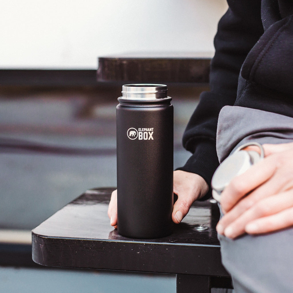 blak 500ml insulated water bottle next to person sat on black bench with lid in her hand.