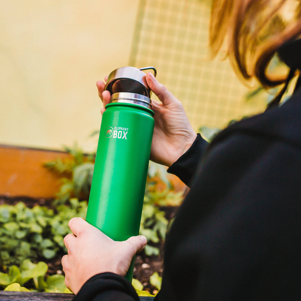 person undoing the steel lid from a green 750ml insulated bottle standing near a raised bed of salad