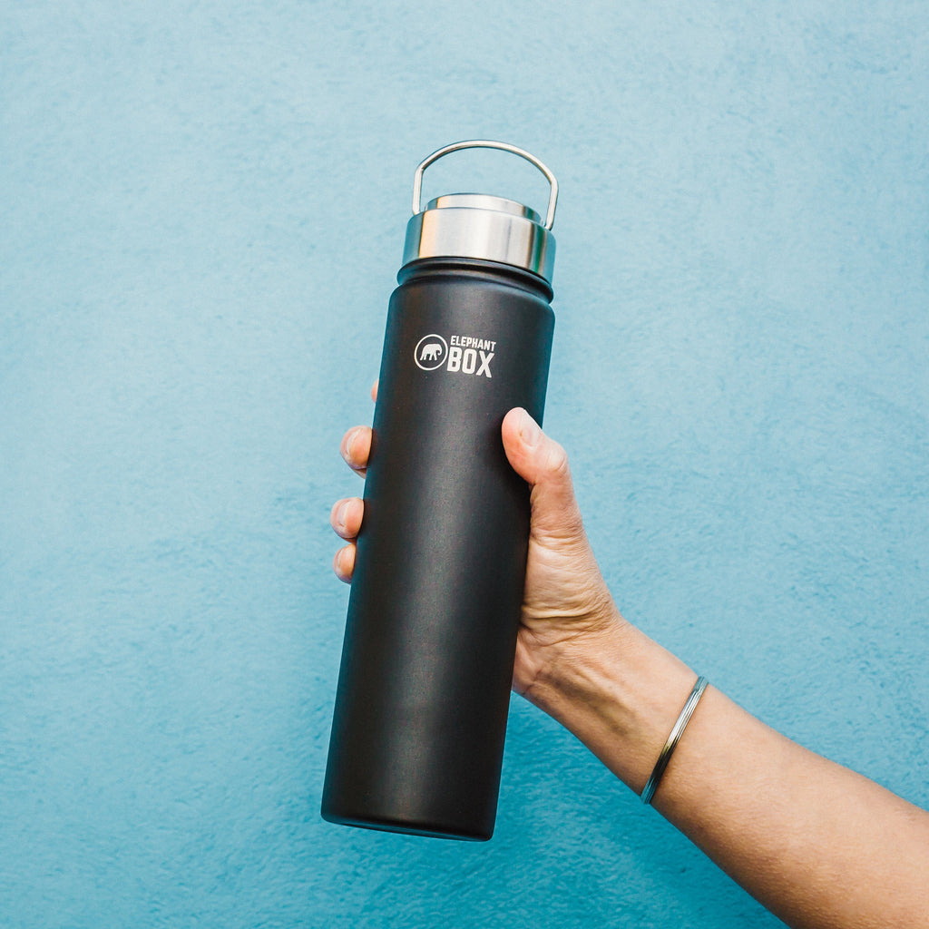 Hand holding a 750ml black insulated water bottle in front of a blue wall.